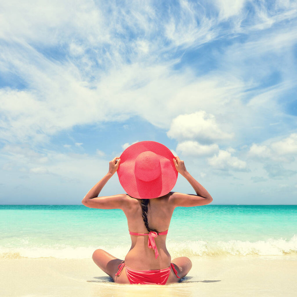 Luxury travel woman relaxing tanning with pink sun hat and bikini sitting on white sand Caribbean beach. Girl tourist on summer holiday at vacation resort. Tropical landscape square for social media. - Fotografie, Obrázek