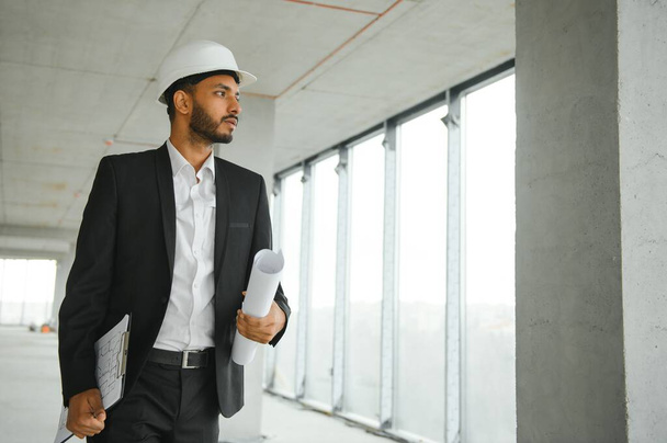 Indian construction site manager standing wearing helmet, thinking at construction site. Portrait of mixed race manual worker or architect - Foto, Imagem