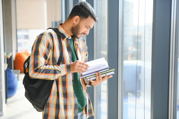 Jonge indiaanse student die boek leest in de universiteitsbibliotheek met boekenplank achter. werken aan opdracht of project. - Foto, afbeelding