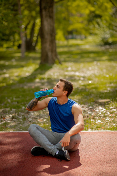 Przystojny mężczyzna siedzi na torze sportowym w parku, zmęczony, po bieganiu w słoneczny dzień. Pije wodę. - Zdjęcie, obraz