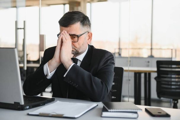 Worried businessman in dark suit sitting at office desk full with books and papers being overloaded with work - Photo, image