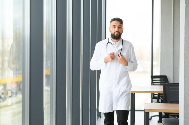 Young 30-aged handsome pleasant Arabic male doctor in white coat, posing at camera indoors - Fotografie, Obrázek