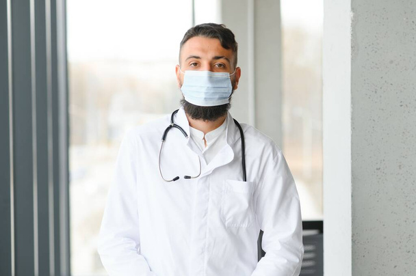 Male pensive thoughtful arab man therapist surgeon wear white uniform medical face mask standing at workplace - Photo, image