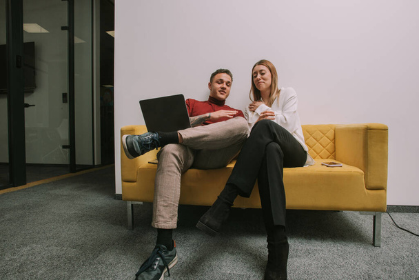 A blonde businesswoman is about to burst out laughing while she is listening to her male colleague talking funny to her while sitting on a sofa at the office - Photo, image