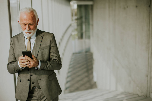 Starszy biznesmen stoi w biurze, skupiony na telefonie komórkowym. Jest ubrany w strój formalny, emanuje pewnością siebie i profesjonalizmem - Zdjęcie, obraz