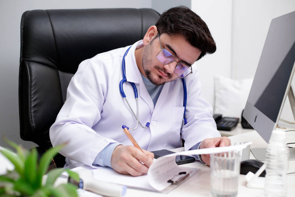 Ein junger Arzt sitzt in der Klinik und schreibt mit Stift etwas auf einen Notizblock. - Foto, Bild