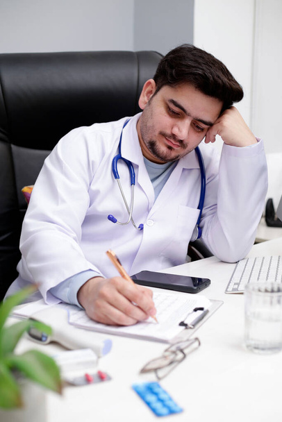 Un médico joven está sentado en la clínica y escribiendo algo en papel con bolígrafo. - Foto, Imagen