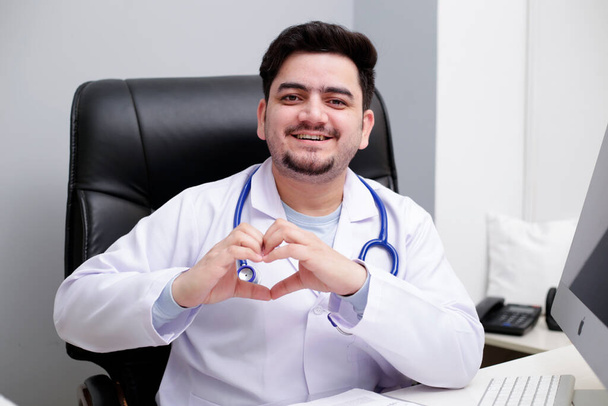Ένας νεαρός γιατρός κάθεται στην κλινική κάνοντας καρδιά με το χέρι και χαμογελώντας. - Φωτογραφία, εικόνα
