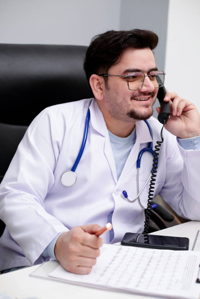 Een jonge dokter zit in de kliniek te telefoneren en te glimlachen.. - Foto, afbeelding