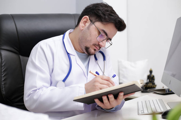 Ein junger Arzt sitzt in der Klinik und schreibt mit Stift etwas ins Tagebuch. - Foto, Bild