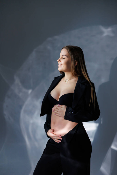 Portrait of a young pregnant woman in a black classic suit. A pregnant woman on a gray background hugs her belly. - Foto, afbeelding