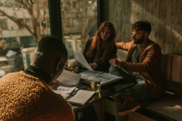 Visszapillantás kép fekete üzletemberről, aki pénzügyi statisztikákat és csapatmunka eredményeket olvas, miközben fehér kollégái együtt dolgoznak az asztal másik oldalán - Fotó, kép