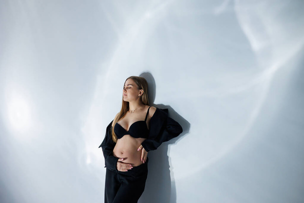 Χαριτωμένη έγκυος στο στούντιο. Έγκυος γυναίκα με μαύρο κοστούμι. Ευτυχισμένη έγκυος γυναίκα - έγκυος φωτογραφία της όμορφης νεαρής μέλλουσας μητέρας σε γκρι φόντο, φωτογραφία στούντιο - Φωτογραφία, εικόνα
