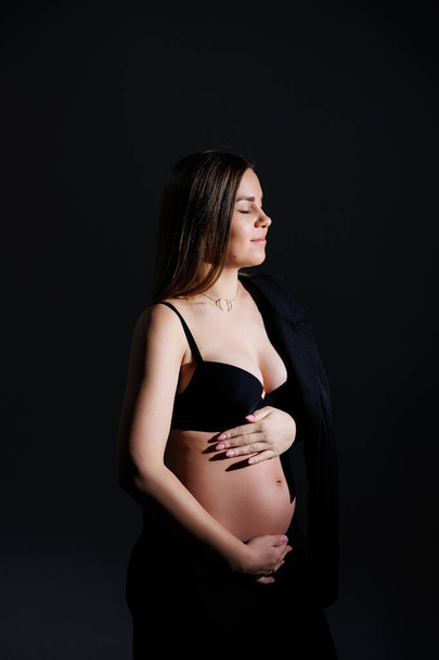 Κομψή έγκυος γυναίκα. Όμορφη έγκυος κορίτσι με μακριά μαλλιά σε μαύρο κοστούμι σε γκρι φόντο, έννοια της ευτυχούς εγκυμοσύνης και της οικογένειας. - Φωτογραφία, εικόνα