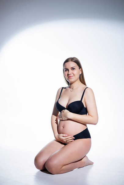 Cute pregnant woman with long hair in black underwear is sitting on a white background. Women's underwear for pregnant women. - Photo, Image