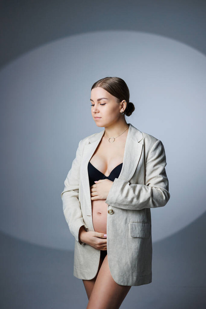 V ateliéru stojí štíhlá těhotná žena v dlouhém saku a spodním prádle na bílém pozadí a drží si ruku na břiše. Dámské spodní prádlo pro těhotné ženy - Fotografie, Obrázek