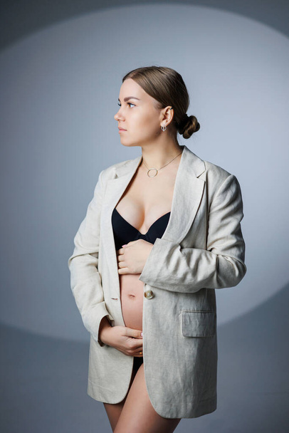 Egy vékony, terhes nő hosszú kabátban és alsóneműben áll a stúdióban fehér háttérben, a kezét a hasán tartva. Női fehérnemű terhes nők számára - Fotó, kép