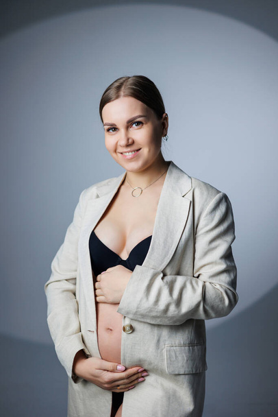 Ohut raskaana oleva nainen, jolla on pitkä takki ja alusvaatteet, seisoo studiossa valkoisella taustalla pitäen kätensä vatsallaan. Naisten alusvaatteet raskaana oleville naisille - Valokuva, kuva