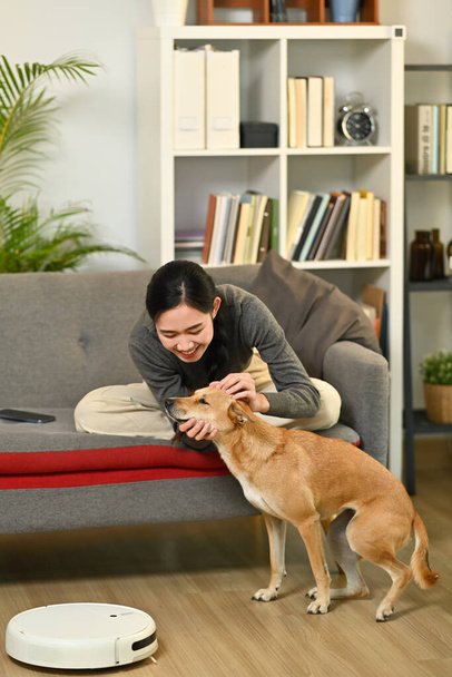 Νεαρή γυναίκα και σκύλος κάθονται στο σαλόνι με ρομποτική ηλεκτρική σκούπα καθαρίζοντας το πάτωμα. Έξυπνο σπίτι και αυτόματη έννοια καθαρισμού. - Φωτογραφία, εικόνα