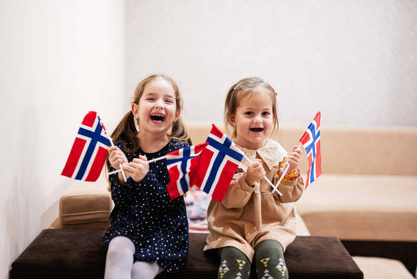 二人の姉妹はノルウェー国旗を手に自宅のソファに座っている。ノルウェーの国旗を持つ子供たち . - 写真・画像