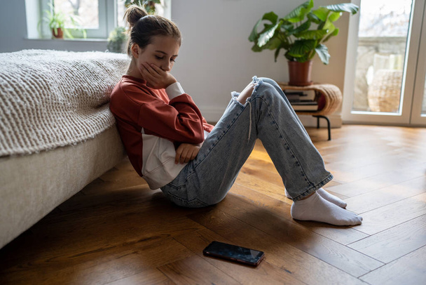 Obrażona, nieszczęśliwa nastolatka, patrząca na telefon, siedzi w domu na podłodze. Smutny troskliwy uczennica przeżywa depresję po zerwaniu z ukochanym chłopcem lub bolesne social media post - Zdjęcie, obraz