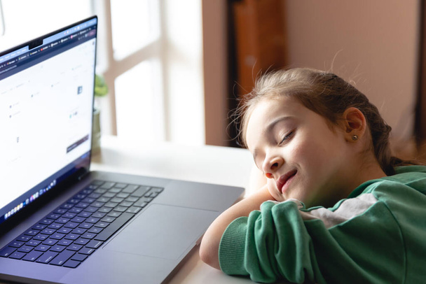 小さな女の子がテーブルの上のノートパソコンの前で寝ていると、疲れた子供がレッスン中に眠ってしまった. - 写真・画像