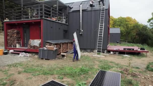 Technikus telepítése napelem rendszer tetején ház. Egy sisakos férfi fotovoltaikus napmodult szállít a szabadban. Az alternatív és megújuló energia fogalma. - Felvétel, videó