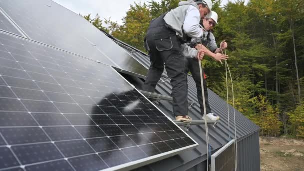 Mannen die zonnepanelen installeren op het dak van huis. Elektriciens in helmen tillen fotovoltaïsche zonnemodule met behulp van touwen buiten. Begrip "alternatieve en hernieuwbare energie". - Video