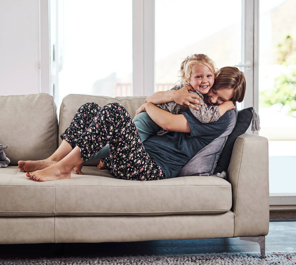 Эта малышка весь мой мир. Полный портрет привлекательной молодой матери, сидящей на диване и обнимающей дочь дома - Фото, изображение
