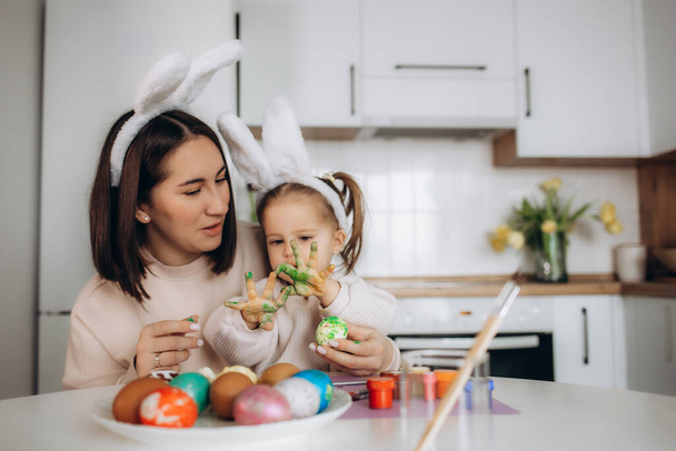 Mother and her daughter with painting eggs. Happy family celebrate Easter. Cute little child girl wearing bunny ears. - Photo, Image