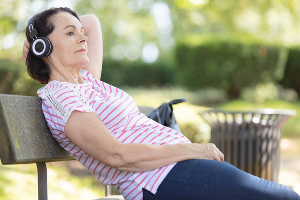 mature woman sat on bench wearing headphones - Фото, изображение