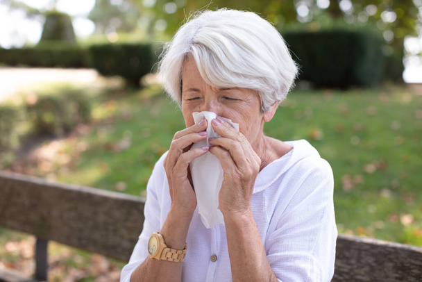 senior woman with tissue having flu or allergy at park - Foto, Imagen