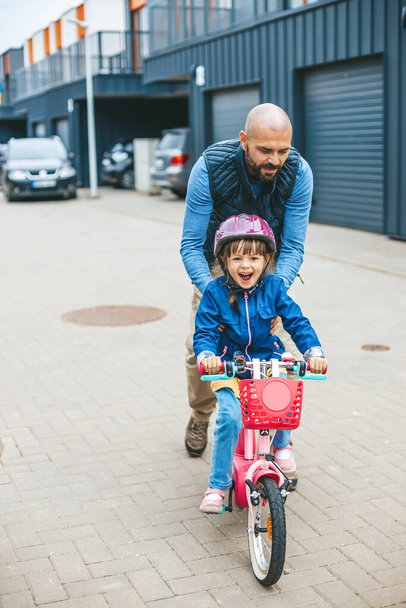 Happy girl riding a bike with her father's hepl. Father teaching his little daughter to ride a bicycle. Family outdoors activities together. - Foto, imagen