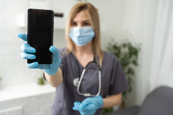 Portrait of young female doctor holding mobile phone and show winning action, with stethoscope and gloves, health mobile application concept. - Zdjęcie, obraz