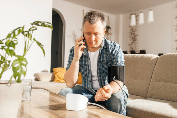 A man measures blood pressure at home with a blood pressure cuff and a tonometer. A man calls a doctor on a mobile phone. Health check at home. Consultations with a doctor online. - Photo, Image