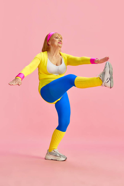 Sportive middle-aged, mature woman in colorful sportswear training, rising legs to hands, posing against pink studio background. Concept of sportive lifestyle, retirement, health care, wellness. Ad - Photo, Image