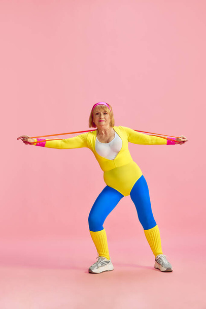 Beautiful mature, senior woman in colorful sportswear training with fitness equipment, posing against pink studio background. Concept of sportive lifestyle, retirement, health care, wellness. Ad - Photo, image