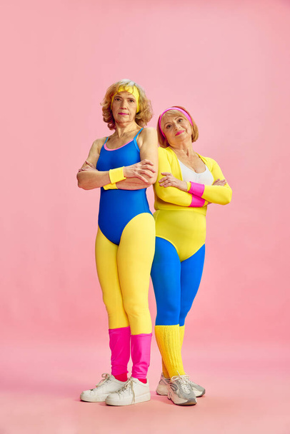 Two senior elderly woman in colorful sportswear posing against pink studio background. Fitness. Keeping fit. Concept of sportive lifestyle, retirement, health care, wellness. Ad - Фото, зображення
