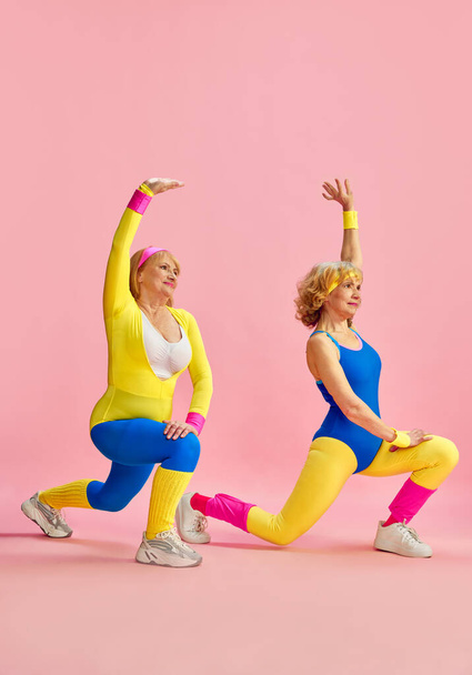 Sportive, beautiful, elderly woman in colorful sportswear training, doing squats and stretching exercises against pink background. Concept of sportive lifestyle, retirement, health care, wellness. Ad - Zdjęcie, obraz