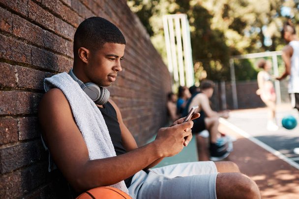Lets see who wants to play. a sporty young man using a cellphone while taking a break after a game of basketball - Foto, afbeelding