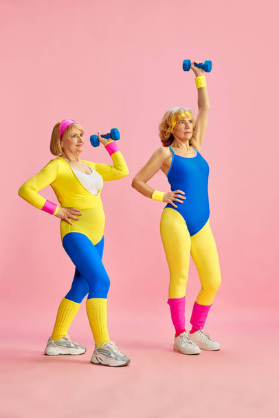 Two senior middle-aged sportive woman in colorful sportswear training, lifting dumbbells against pink studio background. Concept of sportive lifestyle, retirement, health care, wellness. Ad - Photo, image