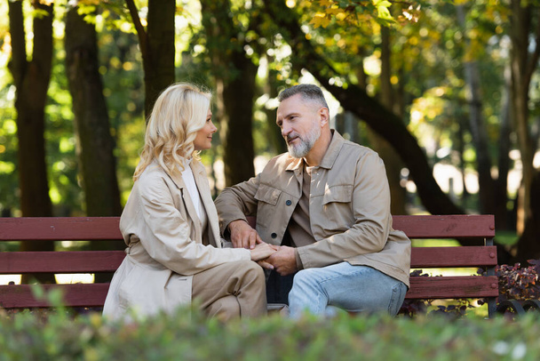 Mature man talking and holding hand of blonde wife while sitting on bench in park  - Foto, imagen