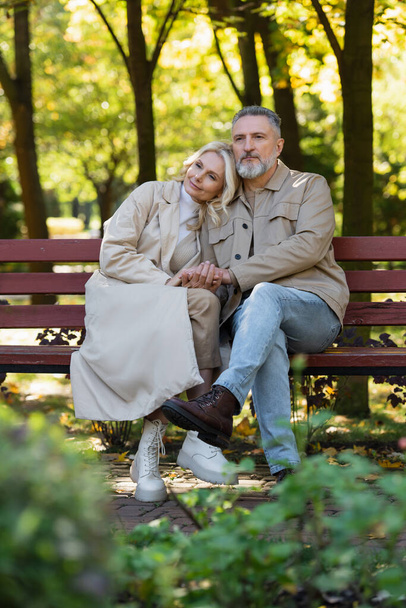 Femme blonde tenant la main d'un mari mature assis sur un banc dans un parc de printemps  - Photo, image