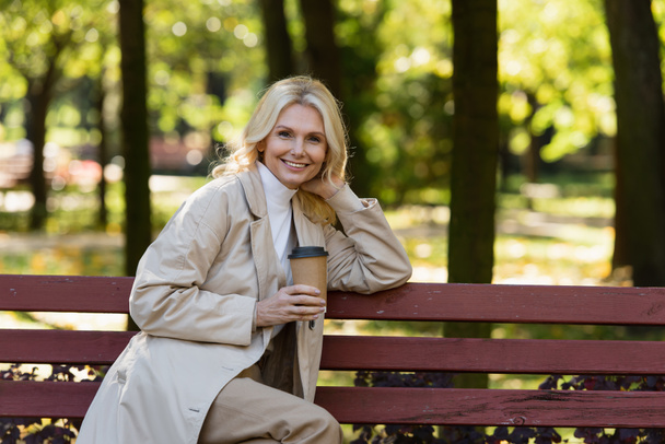 Vidám, érett nő ballonkabátban, kezében kávéval, és ül a padon a parkban.  - Fotó, kép