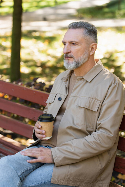 Dojrzały mężczyzna w stroju wiosna trzymając kawę, aby przejść podczas siedzenia na ławce w parku  - Zdjęcie, obraz