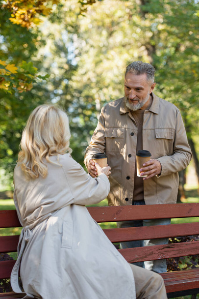 Mature husband giving coffee to go to blonde wife in trench coat in park - Photo, Image