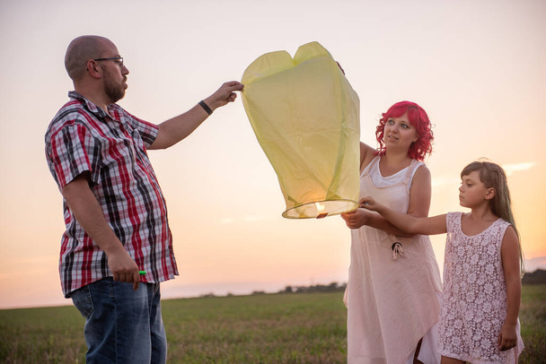 Diversity family, mother father daughter light a yellow paper lantern in the sky at sunset in the field. Teamwork. Making wishes. Concept of hope, faith, travel. Lifestyle happiness. Copy space - Фото, изображение