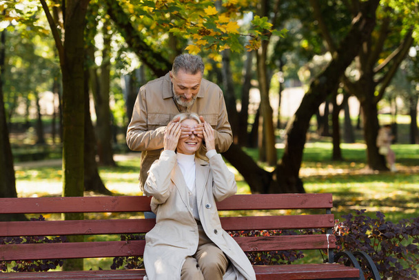 Uśmiechnięty mężczyzna zakrywający oczy żony siedzącej na ławce w parku  - Zdjęcie, obraz