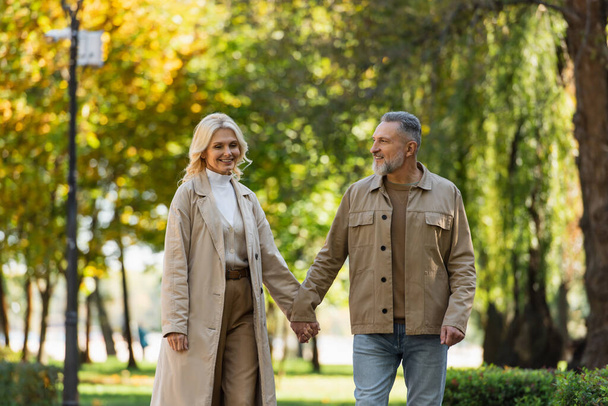 Happy mature man holding hand of blonde wife in trench coat in park  - Фото, зображення