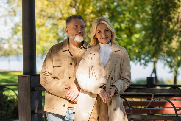 Smiling mature couple standing near bridge in spring park  - Fotografie, Obrázek